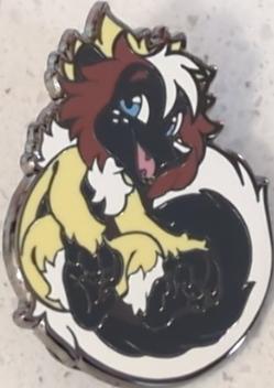A pin depicting Ezra's furry OC, a non-binary male lemur with blue eyes, messy medium-length brown hair with a shock of white in the middle, brown eyebrows, black fur on his face, ears, belly, hands, and center tail, white fur neck and lower leg ruffs, and white fur on the outer edges of his tail. He is sitting down with his tail swept around one leg, and is smiling open-mouthed, looking friendly and enthusiastic.